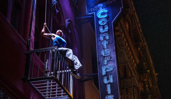 A person with dark skin wearing a blue shirt and gray pants sits on the edge of a large metal cage suspended from the side of a building, hanging from a wire. A blue neon sign reads “San Francisco CounterPulse”