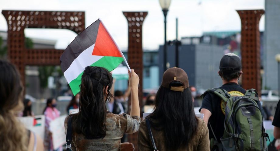 The backs of three people; the person on the left holds up a Palestinian flag.
